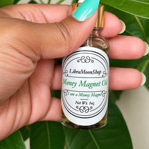Puede incluir: Un frasco cuentagotas de vidrio transparente con cuello dorado y tapa blanca. La etiqueta blanca presenta adornos negros y el texto LibraMoonShop, Money Magnet Oil, I am a Money Magnet y Net Wt. 1oz en tinta negra y verde.