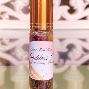 May include: A clear glass roll-on bottle with a gold-coloured cap. The bottle contains a liquid with dried flowers and herbs. The label reads "Libra Moon Shop Goddess Oil I am Divine Feminine."