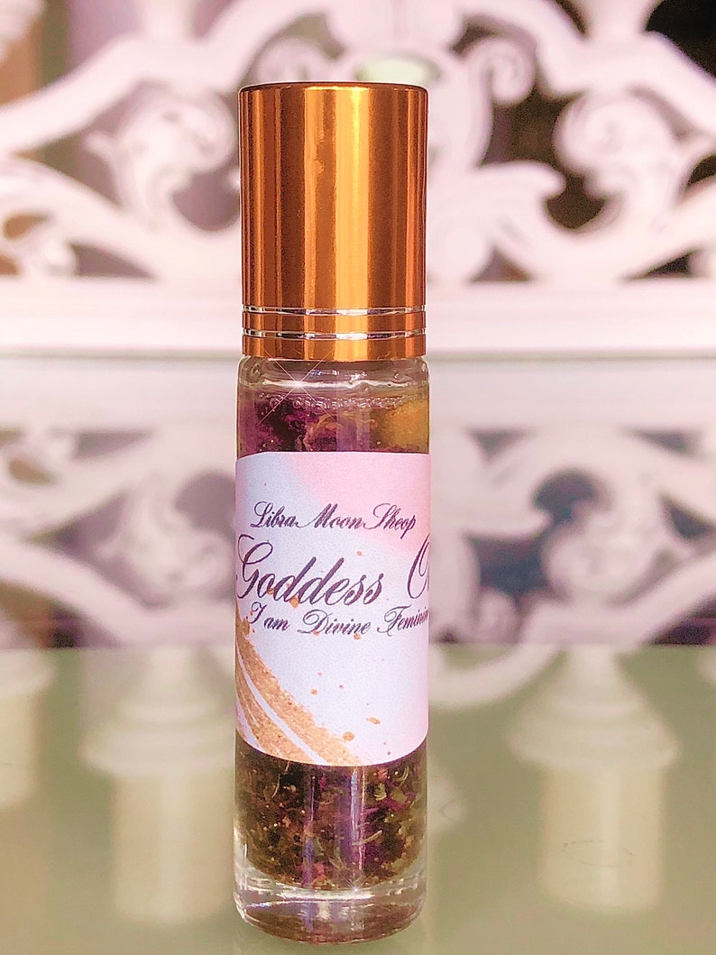 May include: A clear glass roll-on bottle with a gold-coloured cap. The bottle contains a liquid with dried flowers and herbs. The label reads "Libra Moon Shop Goddess Oil I am Divine Feminine."