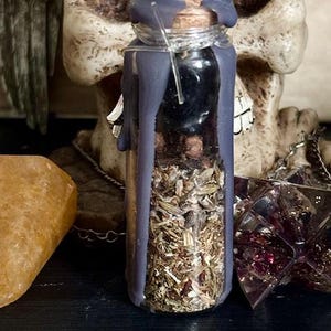 May include: A small glass bottle filled with dried herbs and sealed with a cork stopper. The bottle is covered in a layer of dark purple wax.