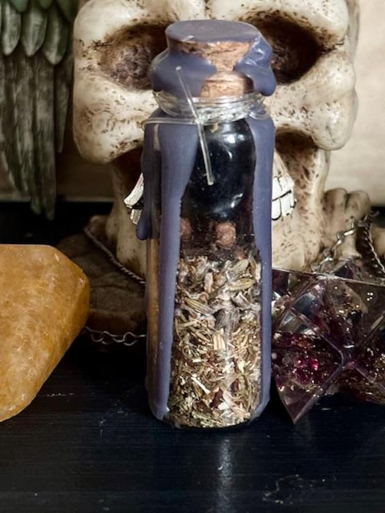 May include: A small glass bottle filled with dried herbs and sealed with a cork stopper. The bottle is covered in a layer of dark purple wax.