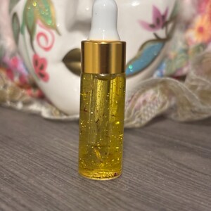 May include: A small, clear glass bottle with a gold-colored cap and a white dropper. The bottle is filled with a yellow liquid containing small, golden particles. A decorative mask is in the background.