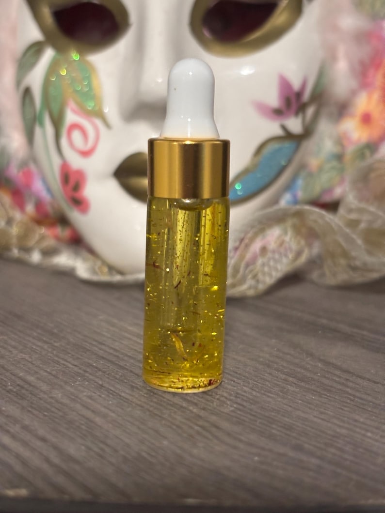 May include: A small, clear glass bottle with a gold-colored cap and a white dropper. The bottle is filled with a yellow liquid containing small, golden particles. A decorative mask is in the background.