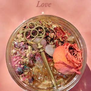 May include: A glass jar filled with honey, herbs, glitter, a rose, a pink crystal, and a gold key. The jar is labeled "Hoodoo Honey Jar Love".