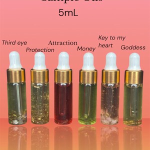 May include: Six small glass sample oil bottles with gold-colored tops and white droppers. The bottles contain various colored liquids and herbs. Text on the image reads "Sample Oils 5mL" and labels each bottle with a different intention: "Third eye", "Protection", "Attraction", "Money", "Key to my heart", and "Goddess".