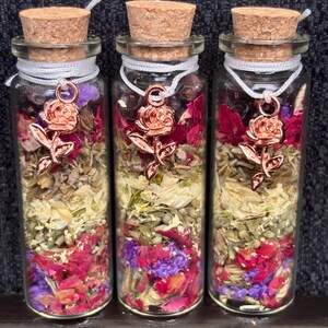Puede incluir: Tres botellas de vidrio llenas de flores y hierbas secas, cada una sellada con un tap&oacute;n de corcho y un dije de rosa. Las botellas contienen una mezcla de flores rojas, moradas y amarillas, con una cuerda blanca alrededor del cuello.