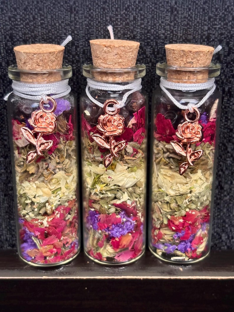 Puede incluir: Tres botellas de vidrio llenas de flores y hierbas secas, cada una sellada con un tap&oacute;n de corcho y un dije de rosa. Las botellas contienen una mezcla de flores rojas, moradas y amarillas, con una cuerda blanca alrededor del cuello.