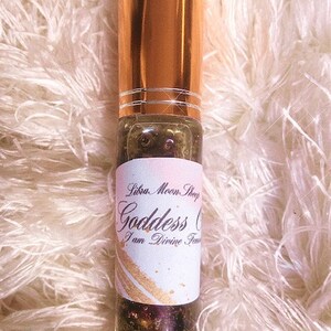 May include: A glass roller bottle with a gold cap and a white label. The label has black text that reads 'Liba Moon Shop Goddess Oil I am Divine Feminine'.