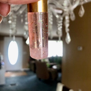 May include: A clear lip gloss tube with a gold cap is held up. The gloss inside is pink with green glitter. The background is blurred, showing a chandelier and a room.