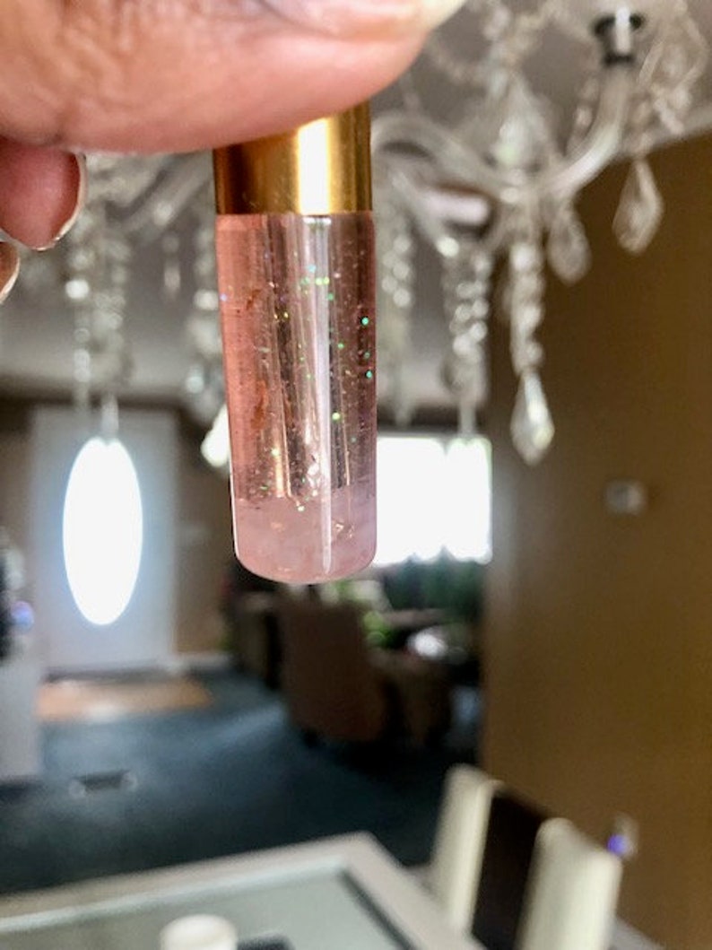 May include: A clear lip gloss tube with a gold cap is held up. The gloss inside is pink with green glitter. The background is blurred, showing a chandelier and a room.