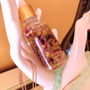 May include: A clear glass bottle with a gold cap and dropper, filled with a liquid containing dried flowers and herbs. The bottle is held in a white ceramic hand, with gold necklaces and a beaded bracelet in the background.