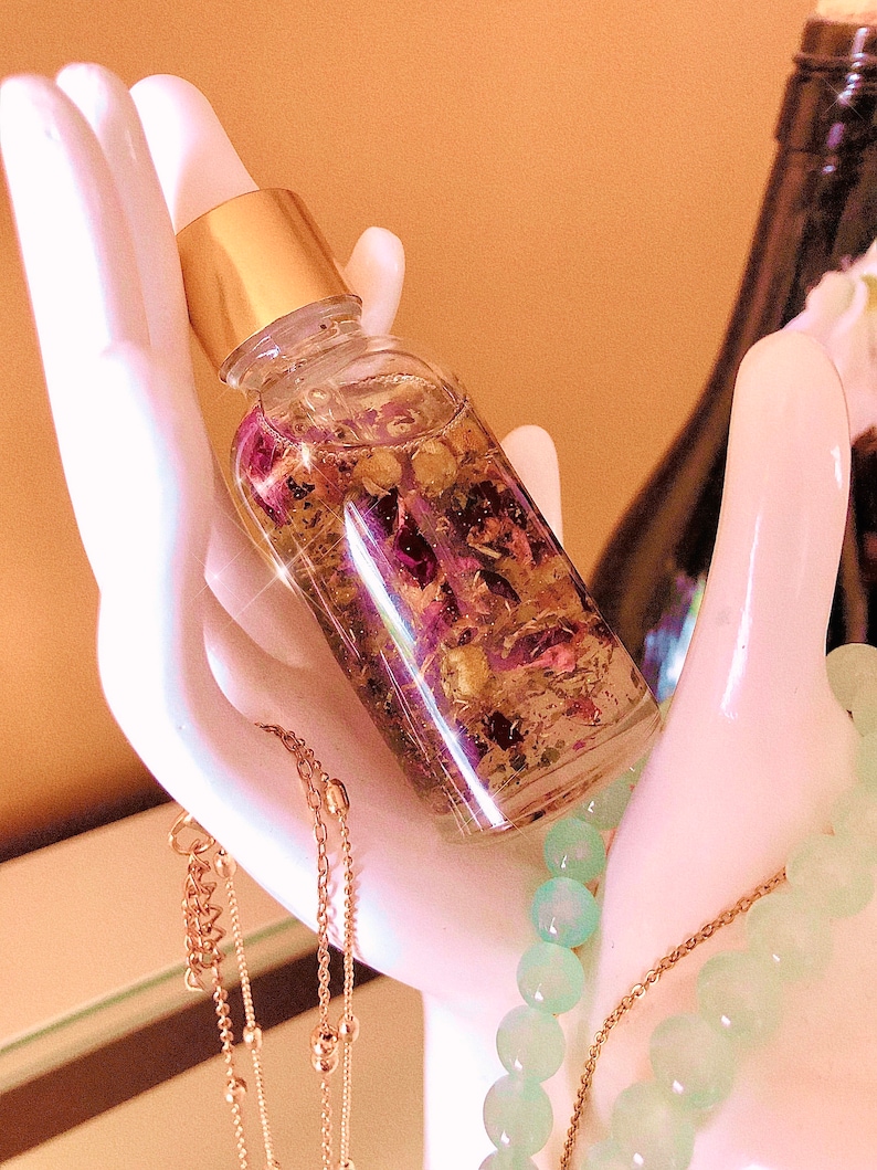 May include: A clear glass bottle with a gold cap and dropper, filled with a liquid containing dried flowers and herbs. The bottle is held in a white ceramic hand, with gold necklaces and a beaded bracelet in the background.