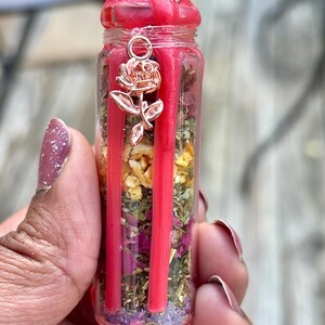 Puede incluir: Una peque&ntilde;a botella de vidrio llena de hierbas secas, flores y cera roja. La botella tiene un dije en forma de rosa colgando de la parte superior. La parte superior de la botella est&aacute; sellada con cera roja.