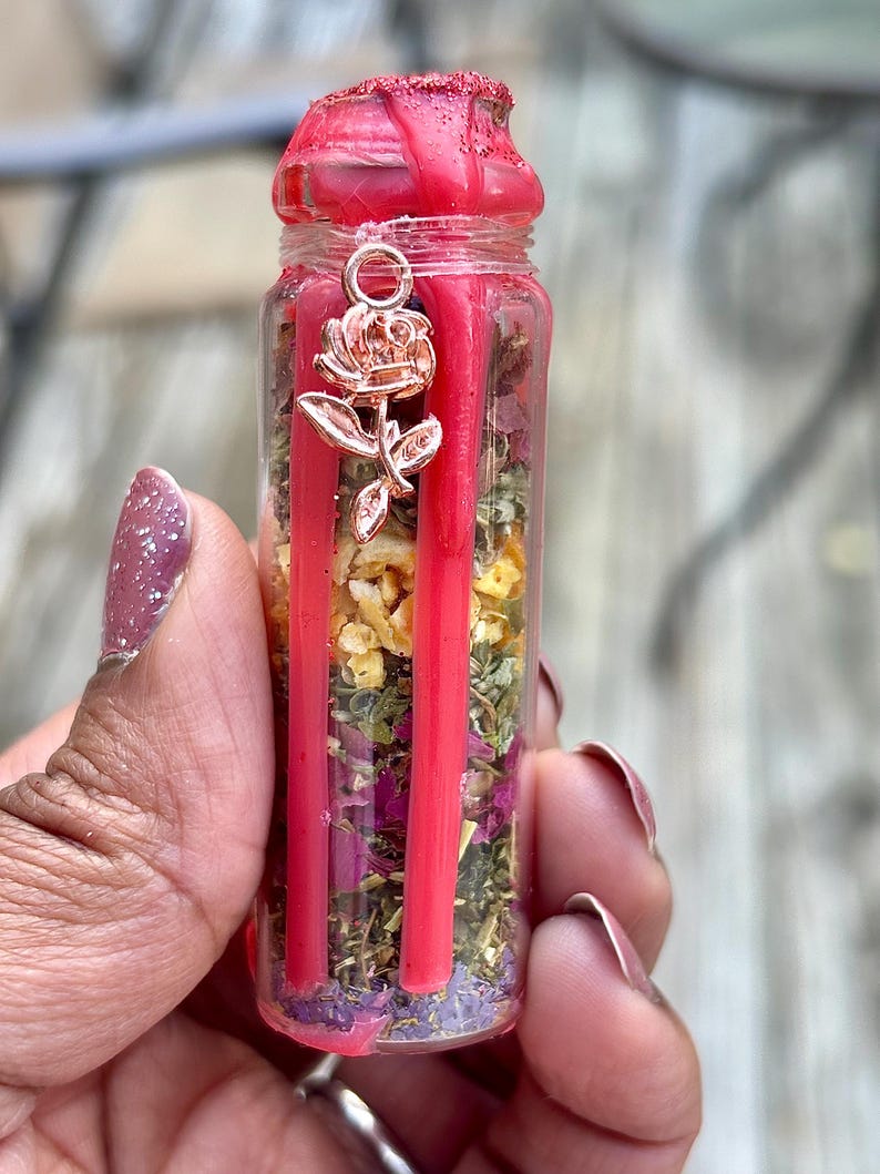 Puede incluir: Una peque&ntilde;a botella de vidrio llena de hierbas secas, flores y cera roja. La botella tiene un dije en forma de rosa colgando de la parte superior. La parte superior de la botella est&aacute; sellada con cera roja.