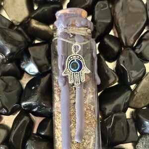 May include: A small glass bottle filled with dried herbs and topped with a purple wax seal. A silver charm with a blue evil eye hangs from the wax seal.