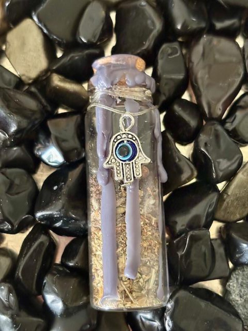 May include: A small glass bottle filled with dried herbs and topped with a purple wax seal. A silver charm with a blue evil eye hangs from the wax seal.