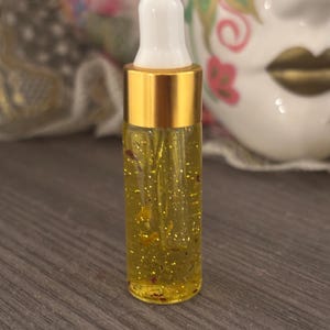 May include: A small glass bottle filled with a yellow liquid containing gold flecks and small red particles. The bottle has a gold metal cap and a white dropper. The background is blurred, but includes a decorative mask.