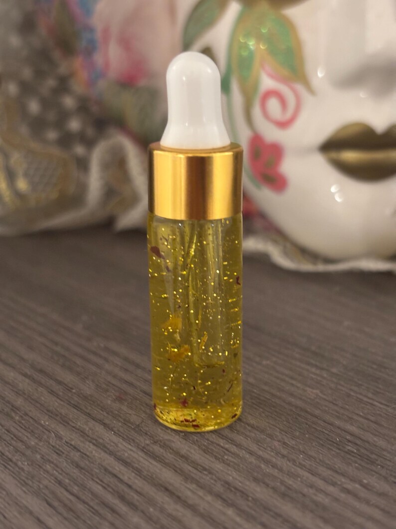 May include: A small glass bottle filled with a yellow liquid containing gold flecks and small red particles. The bottle has a gold metal cap and a white dropper. The background is blurred, but includes a decorative mask.
