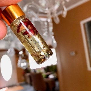 May include: A clear glass roller bottle with a gold cap, filled with a golden liquid containing dried rose petals and gold flakes. The bottle is held up against a blurred background.
