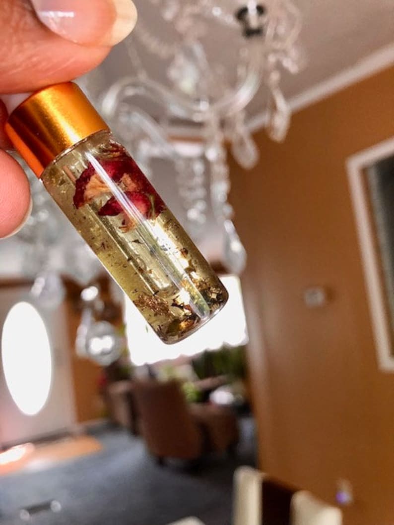 May include: A clear glass roller bottle with a gold cap, filled with a golden liquid containing dried rose petals and gold flakes. The bottle is held up against a blurred background.