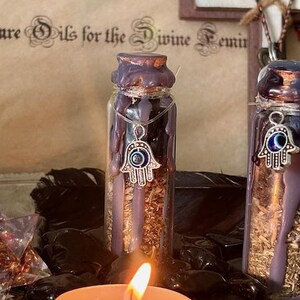 May include: Two small glass bottles with black wax and a silver hamsa hand charm with an evil eye design. The bottles are filled with dried herbs and are sitting on a black surface. A lit candle is in the foreground.