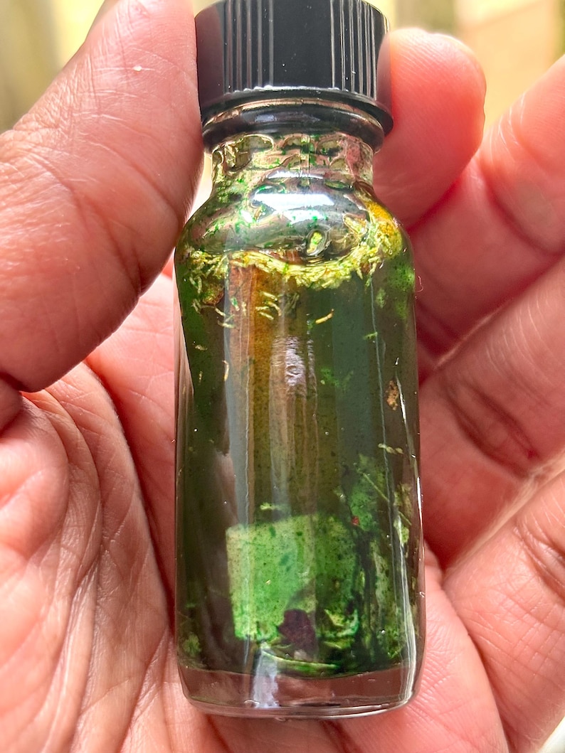 Pode incluir: Uma pequena garrafa de vidro transparente com tampa preta, cheia de l&iacute;quido verde e mat&eacute;ria vegetal. A garrafa &eacute; segurada na m&atilde;o, mostrando o conte&uacute;do. O l&iacute;quido parece ser infundido com ervas ou bot&acirc;nicos.