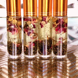 May include: Four clear glass roll-on bottles with gold-coloured caps. Each bottle contains a clear liquid with dried flowers and herbs. The bottles are arranged on a gold and white chevron patterned surface.
