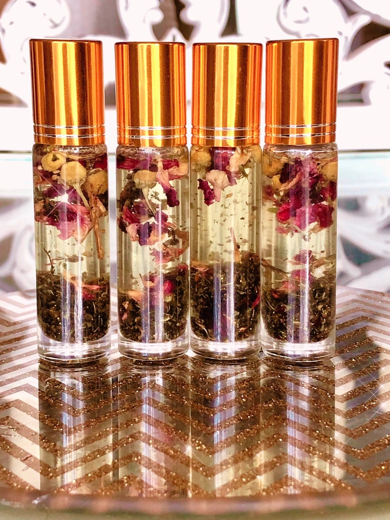 May include: Four clear glass roll-on bottles with gold-coloured caps. Each bottle contains a clear liquid with dried flowers and herbs. The bottles are arranged on a gold and white chevron patterned surface.
