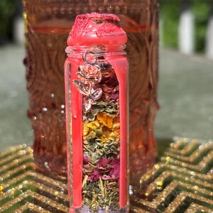 Puede incluir: Un peque&ntilde;o frasco de vidrio lleno de capas de hierbas y flores secas, sellado con cera roja. Un dije de rosa plateada cuelga de la tapa. El frasco se coloca contra un fondo borroso de un frasco m&aacute;s grande y similar y una superficie dorada y brillante.