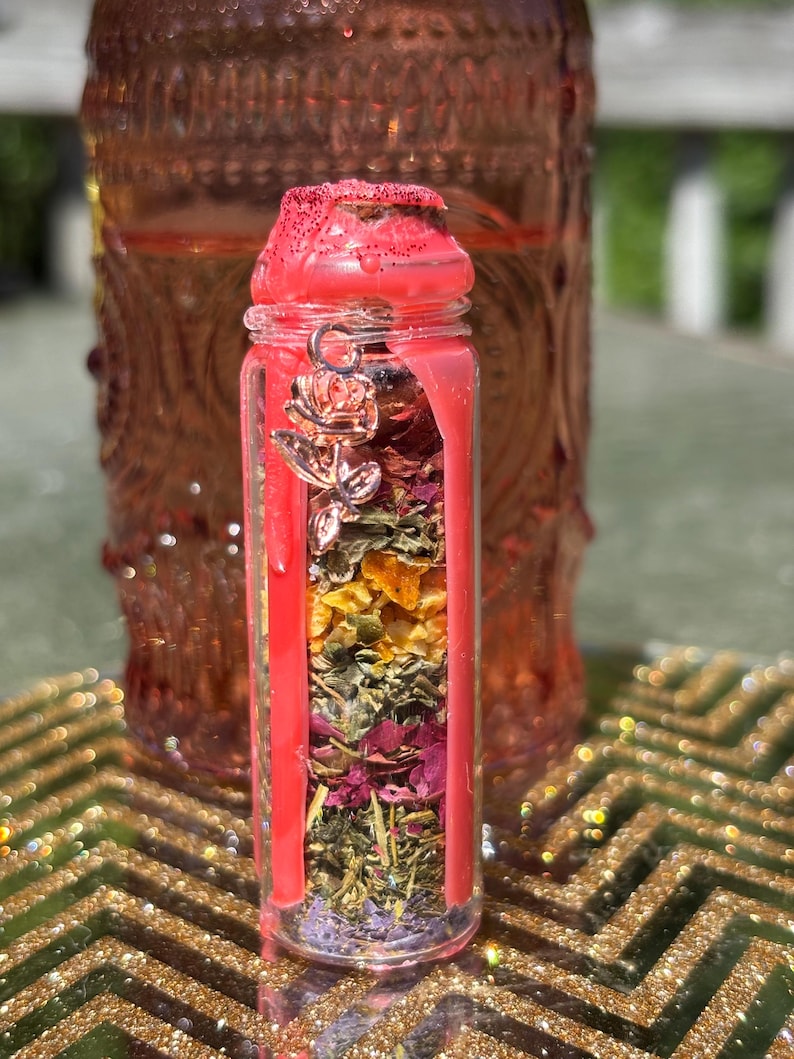 Puede incluir: Un peque&ntilde;o frasco de vidrio lleno de capas de hierbas y flores secas, sellado con cera roja. Un dije de rosa plateada cuelga de la tapa. El frasco se coloca contra un fondo borroso de un frasco m&aacute;s grande y similar y una superficie dorada y brillante.