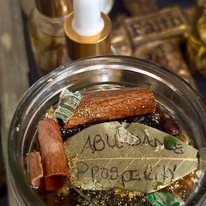 Puede incluir: Un tarro de cristal transparente lleno de líquido, palitos de canela y una hoja de laurel con las palabras "Abundance Prosperity" escritas. También se ven purpurina dorada y pequeños trozos de papel. Dos botellas pequeñas con tapas doradas están en el fondo.