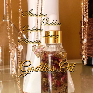 May include: A clear glass bottle of Goddess Oil with gold detailing and a gold cap, filled with dried flowers and herbs. Gold necklaces with charms and pearls hang around the bottle. The words "Attraction, Confidence, Attention, Goddess, Luxury, Goddess Oil" are written on the image.