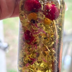 May include: A clear glass bottle filled with a golden liquid, dried flowers, and glitter. The bottle contains red rose buds, yellow chamomile flowers, and small pink petals. The bottle is held up against a blurred background.