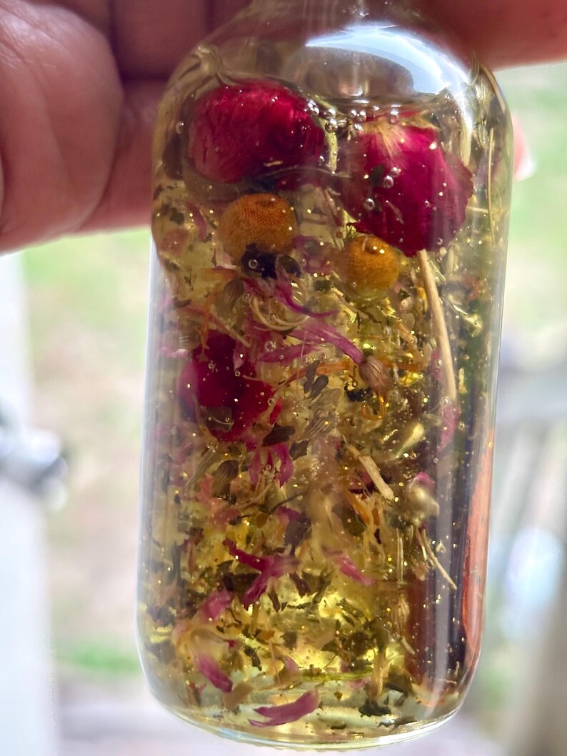 May include: A clear glass bottle filled with a golden liquid, dried flowers, and glitter. The bottle contains red rose buds, yellow chamomile flowers, and small pink petals. The bottle is held up against a blurred background.