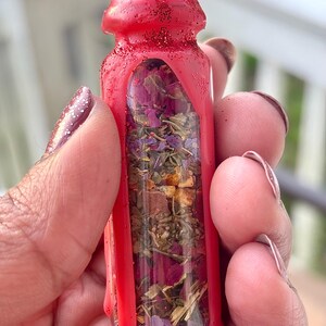 Puede incluir: Una peque&ntilde;a botella de vidrio transparente llena de hierbas y flores secas, encerrada en una carcasa roja vibrante con purpurina. La botella se sostiene en una mano, mostrando el colorido contenido.