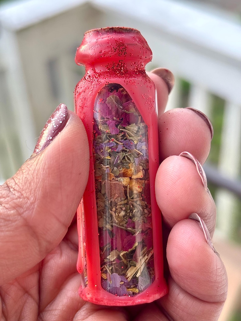 Puede incluir: Una peque&ntilde;a botella de vidrio transparente llena de hierbas y flores secas, encerrada en una carcasa roja vibrante con purpurina. La botella se sostiene en una mano, mostrando el colorido contenido.