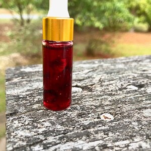 May include: A small glass bottle with a white dropper top and a gold-colored cap. The bottle is filled with a dark red liquid, possibly a serum or essential oil. The background is blurred, with green foliage.