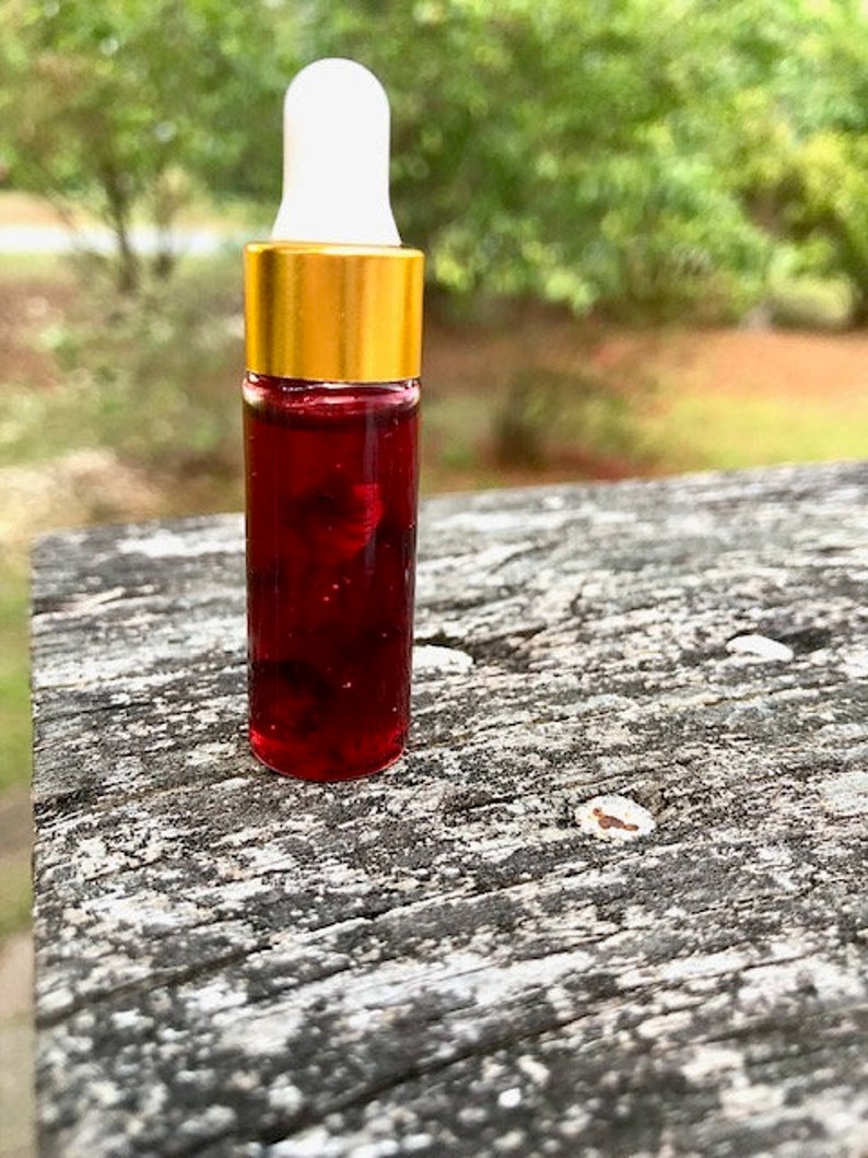 May include: A small glass bottle with a white dropper top and a gold-colored cap. The bottle is filled with a dark red liquid, possibly a serum or essential oil. The background is blurred, with green foliage.