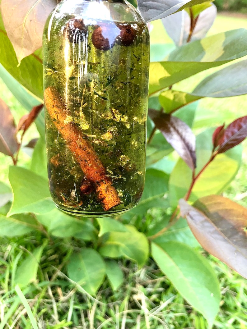 May include: A clear glass bottle with a brown cinnamon stick and green herbs inside. The bottle is filled with a light yellow liquid.