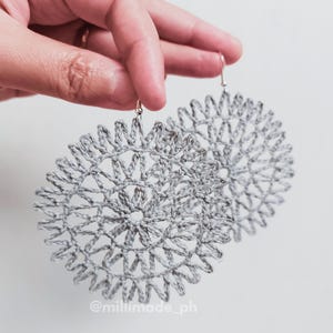May include: A pair of large, gray crocheted earrings. The earrings are circular with a detailed, openwork design. The earrings are held up by a hand against a white background. The text "@milimade_ph" is visible.