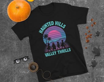 May include: A black short-sleeve t-shirt featuring a graphic print of a sunset over palm trees and silhouettes of four children trick-or-treating. The text reads HAUNTED HILLS and VALLEY THRILLS in a light blue font with a dripping effect.