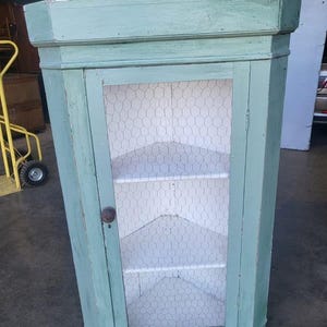 May include: A vintage corner cabinet painted in a light blue shade. The cabinet has a glass door with a wire mesh insert and two shelves. The cabinet features a simple wooden knob.
