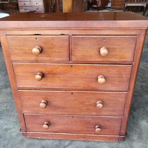 May include: A wooden chest of drawers in a warm brown hue. It has five drawers, each with a round wooden knob and keyhole. The chest is rectangular and designed for storage, with a height of approximately 100 cm.