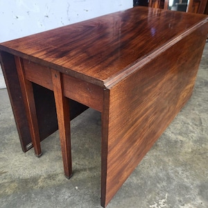 May include: A rectangular wooden drop-leaf table with a polished dark brown finish and visible wood grain. The table features a central fixed section and two hinged side leaves that hang vertically, supported by a sturdy wooden frame and legs.