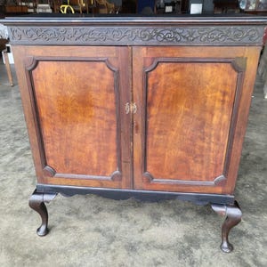 May include: A two-door wooden cabinet with a dark brown finish. The cabinet features ornate carvings along the top edge and decorative panels on the doors. The cabinet rests on four curved legs.