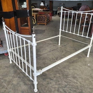May include: White metal bed frame with a headboard and footboard. The frame has a simple design with vertical bars and a curved top.