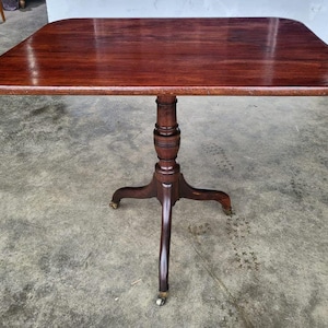 May include: A dark mahogany side table with a rectangular top and rounded corners. The table is supported by a central column and three curved legs, each with a small caster wheel. The table's surface is polished and reflects light.