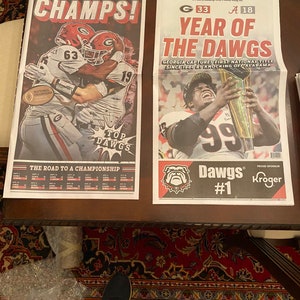 May include: Two front pages of the Atlanta Journal-Constitution newspaper celebrating the University of Georgia Bulldogs' victory in the College Football Playoff National Championship. The headline on the left page reads "CHAMPS!" and the headline on the right page reads "YEAR OF THE DAWGS".