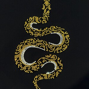 May include: A stylized snake design composed of yellow and white rhinestones on a black background. The snake is coiled, with the head at the top and the tail curving downwards. The rhinestones create a shimmering effect.