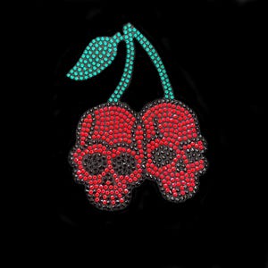 Op de afbeelding: Een met strass versierd ontwerp van twee kersen, elk in de vorm van een schedel, tegen een zwarte achtergrond. De kersen zijn rood met zwarte accenten en de stelen en het blad zijn turquoise. Het ontwerp is waarschijnlijk voor kleding of accessoires.